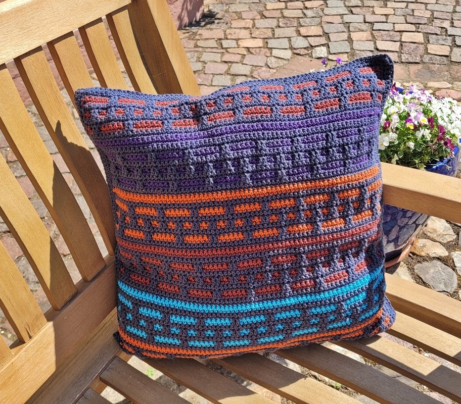 Gehäkeltes quadratisches Kissen mit geometrischem Muster in Dunkelblau, Orange, Violett und Türkis auf einer Holzbank.