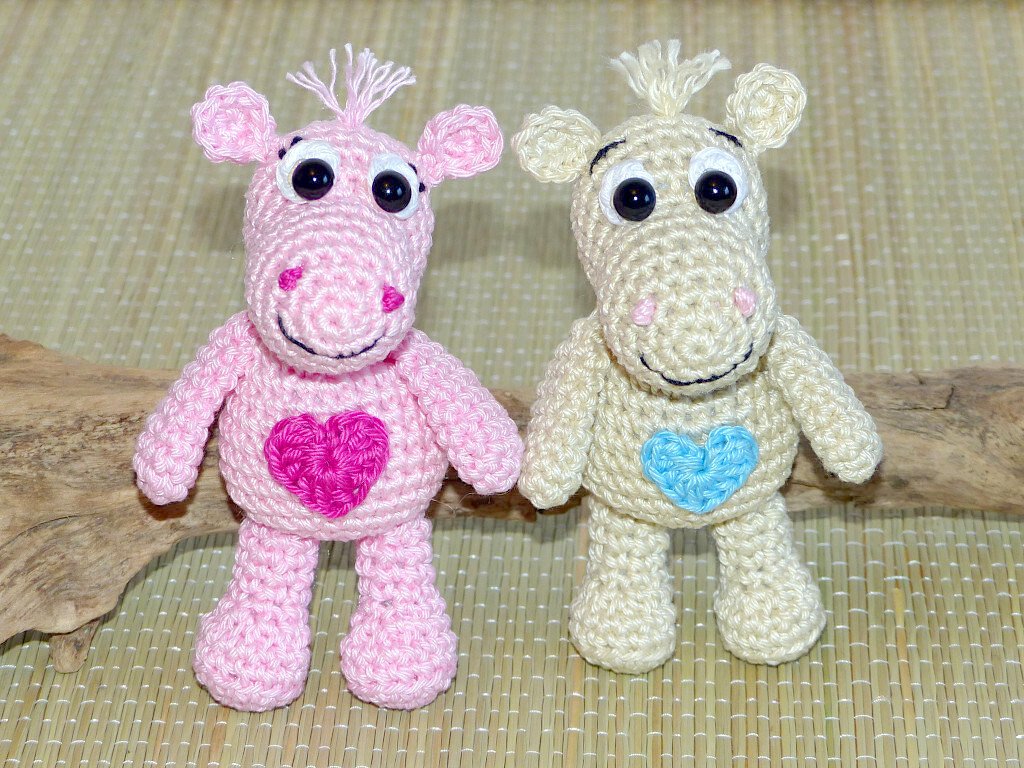 Zwei gehäkelte Amigurumi‑Nilpferde, rosa mit pinkem Herz und beige mit hellblauem Herz, nebeneinander auf einer Matte.