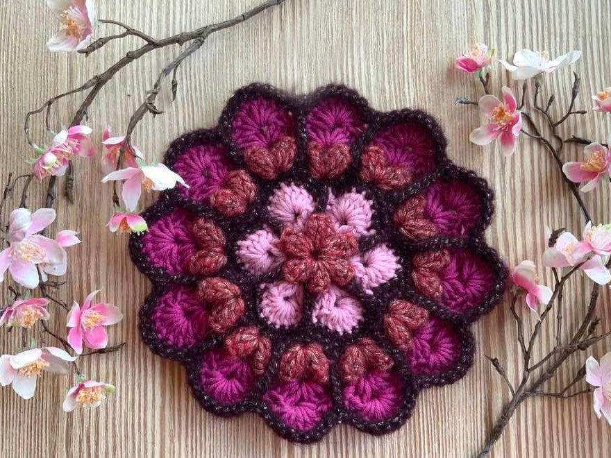 Mehrlagige gehäkelte Blumenapplikation in Rosa- und Beerentönen auf Holzuntergrund