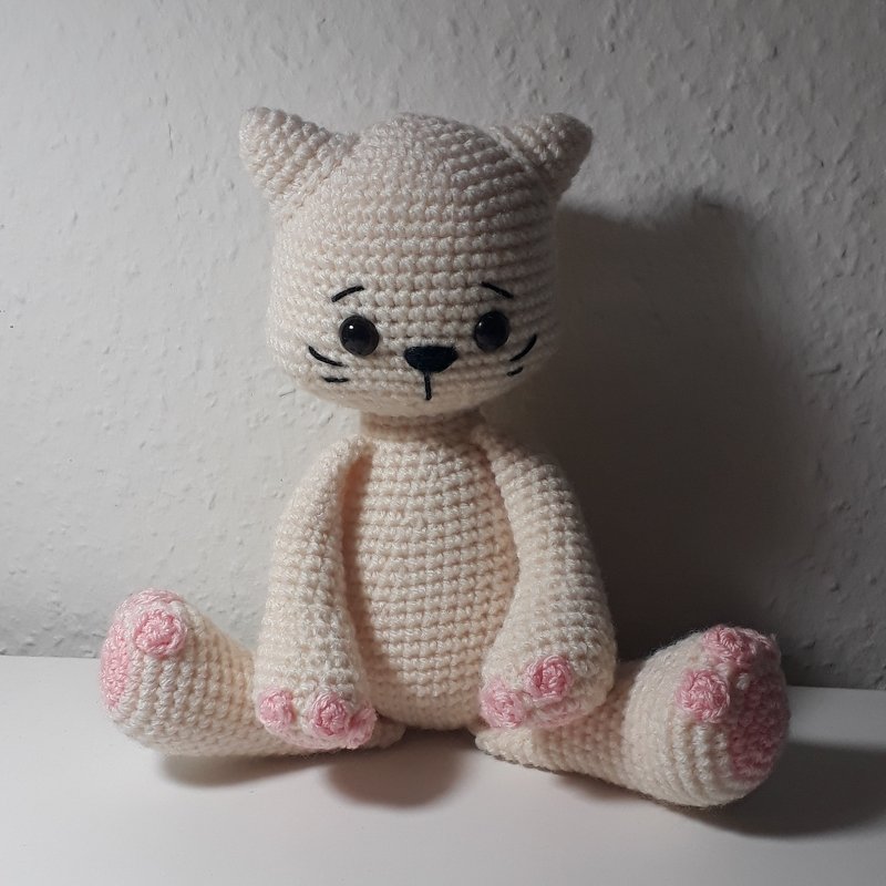 Cremefarbene Amigurumi-Katze mit rosa Pfotenballen, schwarzen Augen und gestickten Schnurrhaaren, sitzend.