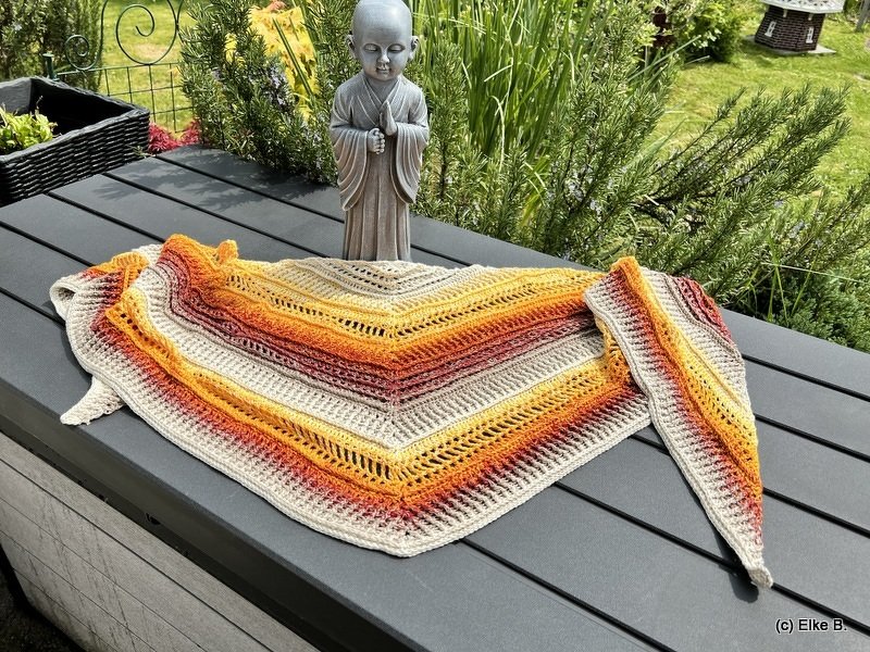 Crocheted triangle shawl in beige, yellow, orange and rust gradient laid flat on a dark slatted table with a small garden statue and greenery behind