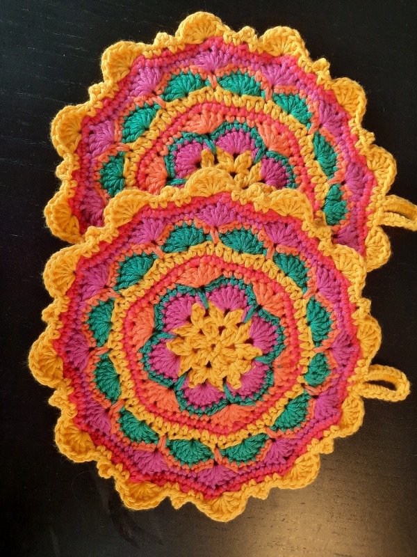 Zwei runde gehäkelte Topflappen in Gelb, Pink, Orange und Grün mit gezacktem Muschelrand und Aufhängeöse