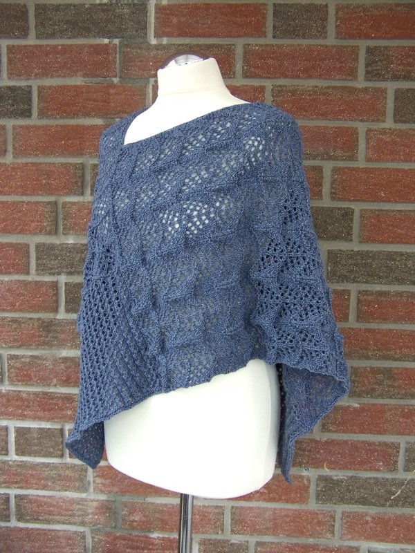 Blauer gestrickter asymmetrischer Poncho mit Lochmuster auf Schneiderpuppe vor Backsteinwand