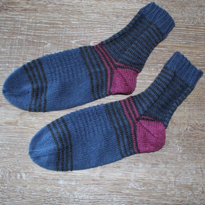 Handgestrickte Damensocken in Dunkelblau mit schwarzen Streifen und magentafarbenen Fersen und Spitzen auf Holz.