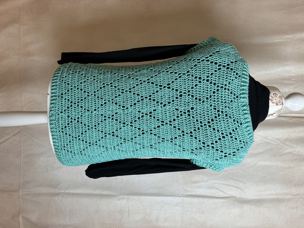 Türkise gehäkelte ärmellose Weste mit Rautenmuster auf Schneiderpuppe über schwarzem Langarmshirt.