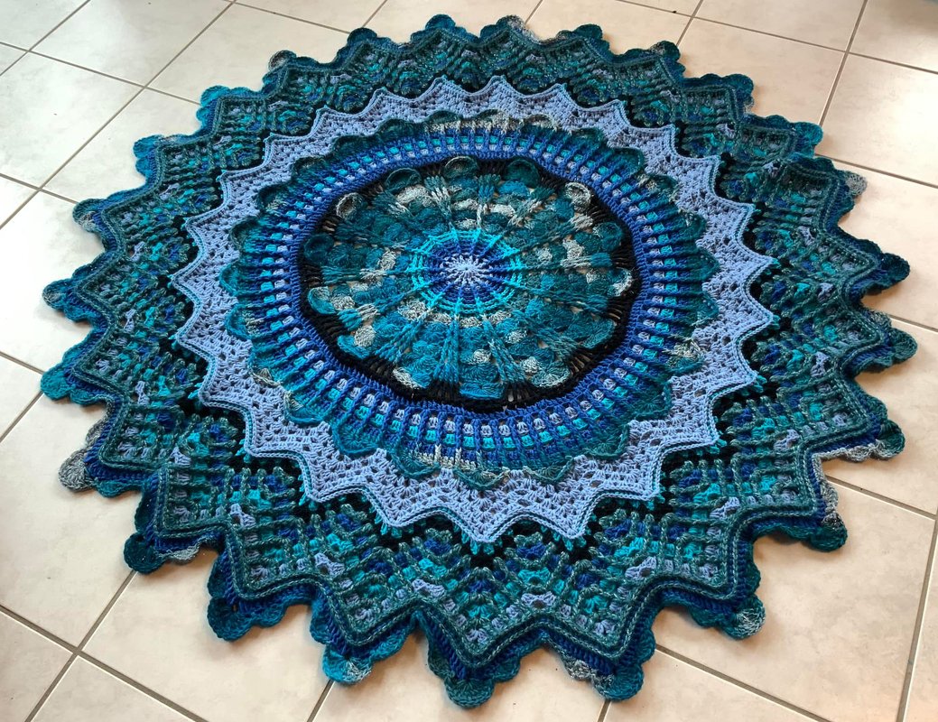 Runde gehäkelte Decke in Blau- und Türkistönen mit komplexem Mandala-Muster und gewelltem Rand auf hellem Boden.