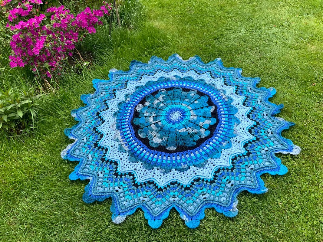 Runde gehäkelte Mandala-Decke in Blau- und Türkistönen auf grünem Rasen neben rosa Blüten.
