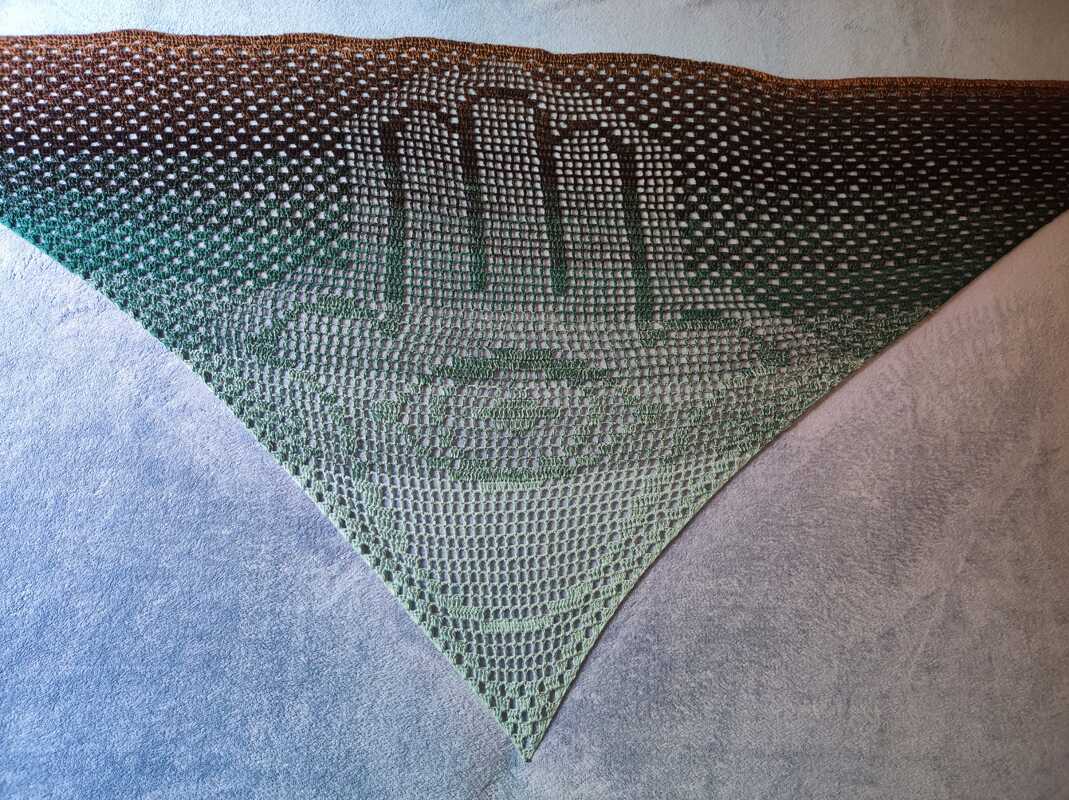 Gehäkeltes Dreieckstuch im Filetmuster mit zentralem Handmotiv und Farbverlauf von Braun zu Grün, ausgebreitet auf hellem Untergrund