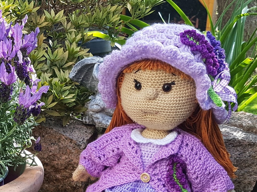 Handgehäkelte Puppe mit rotbraunem Haar, lila Hut und passendem Cardigan neben Lavendel