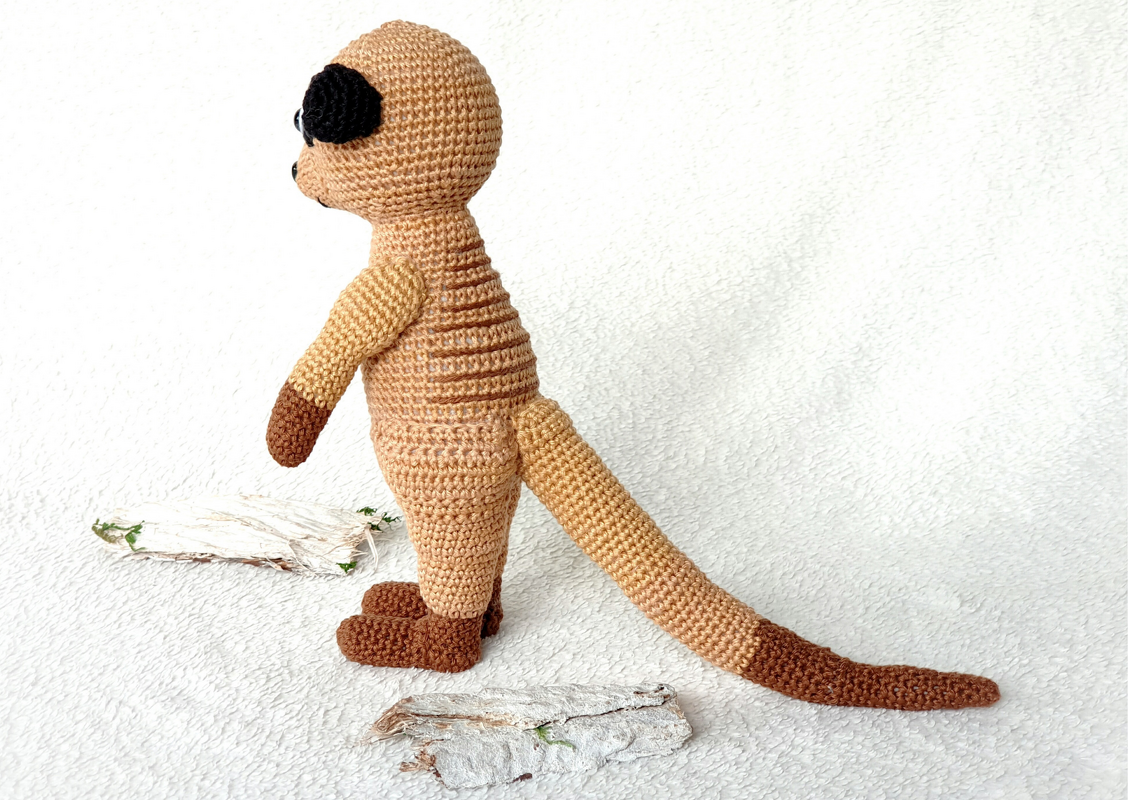 Gehäkeltes Erdmännchen in Beige- und Brauntönen, seitliche Ansicht auf hellem Hintergrund.