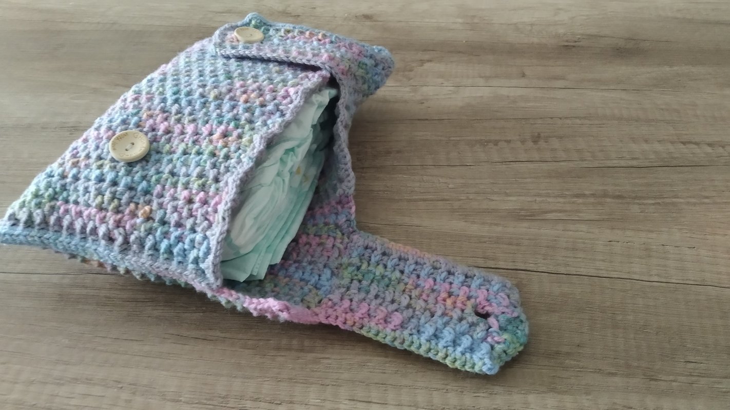 Gehäkelte Wickeltasche in Pastellfarben mit zwei Holzknöpfen, gefüllt mit Windeln auf hellem Holzuntergrund