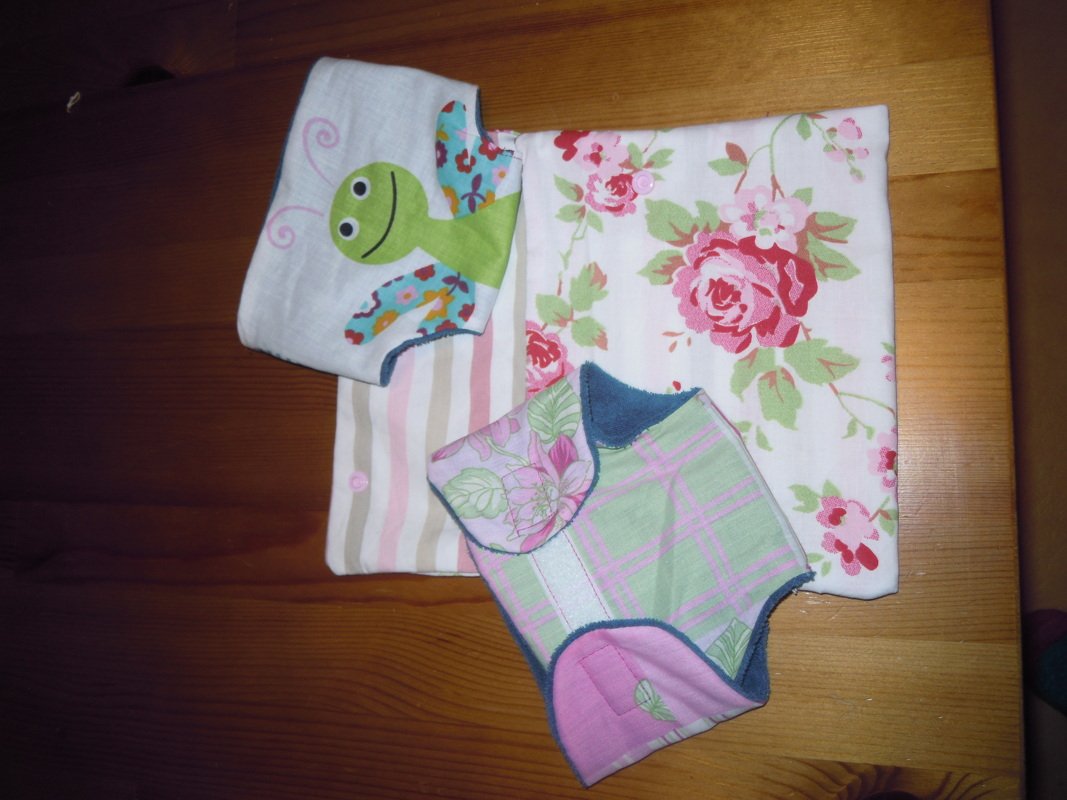 Genähte Baby- und Puppenaccessoires: Lätzchen mit grünem Käfer, gestreifter Stoff, geblümter Stoff und kleine Puppenwindel.