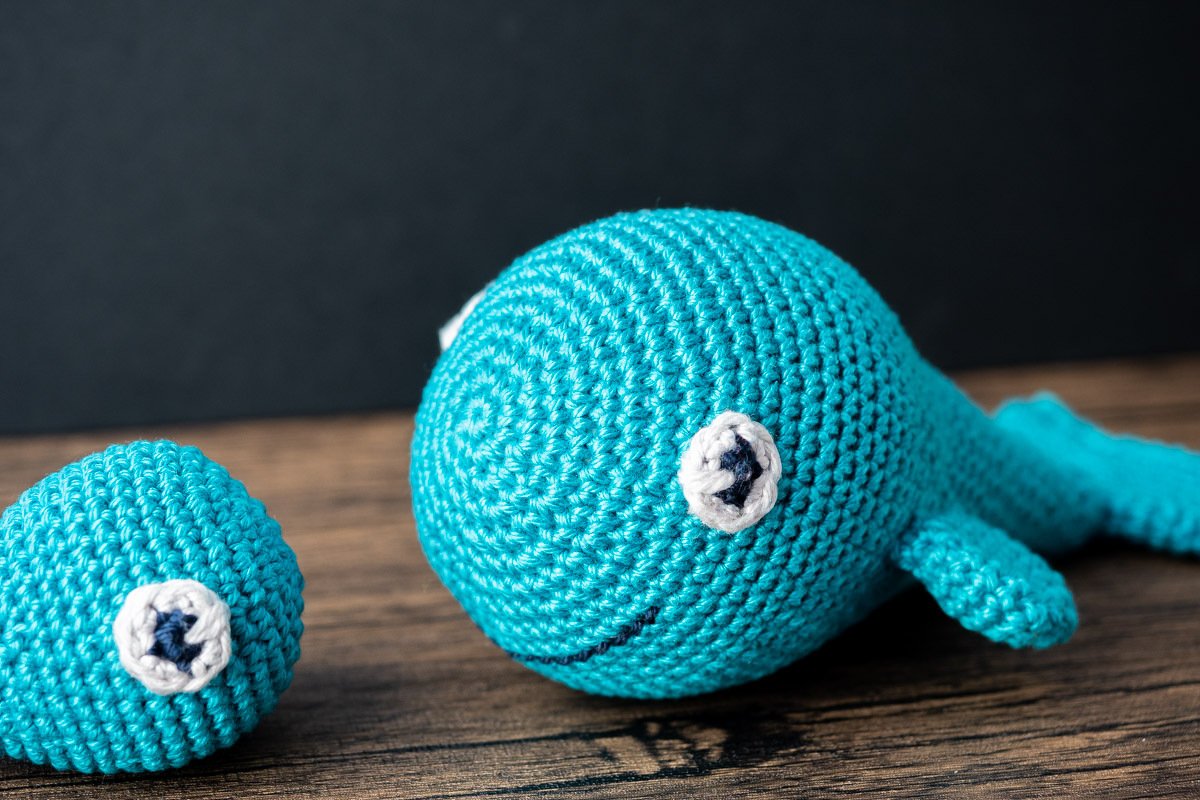 Türkisblauer Amigurumi‑Wal mit lächelndem Mund und kleinem rundem Häkelteil auf Holztisch