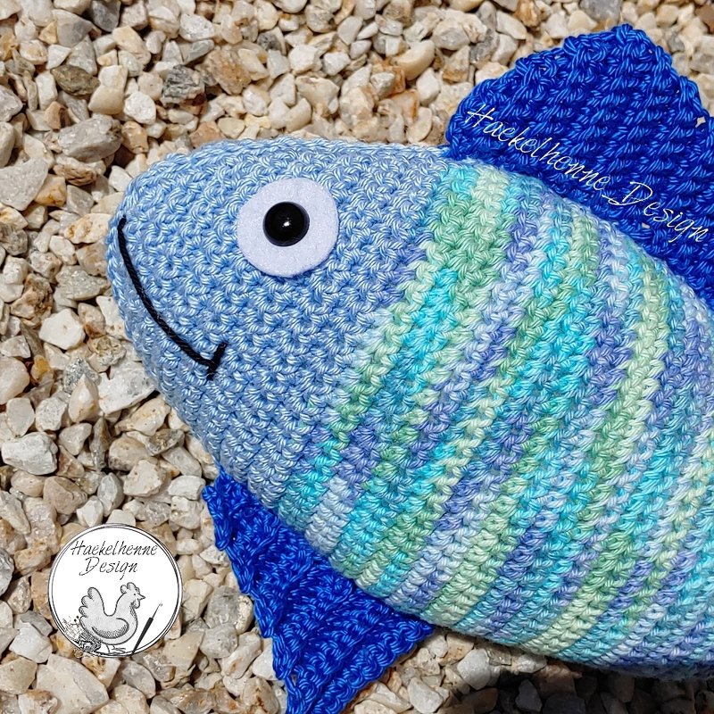 Nahaufnahme eines gehäkelten Fisches mit gestreiftem Körper, Filzauge und dunkelblauen Flossen auf Kies