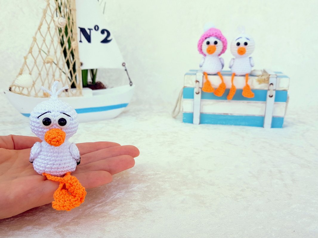 Weiße gehäkelte Möwe mit orangefarbenem Schnabel und Füßen in einer Hand; im Hintergrund zwei weitere Vögel und ein Segelboot.