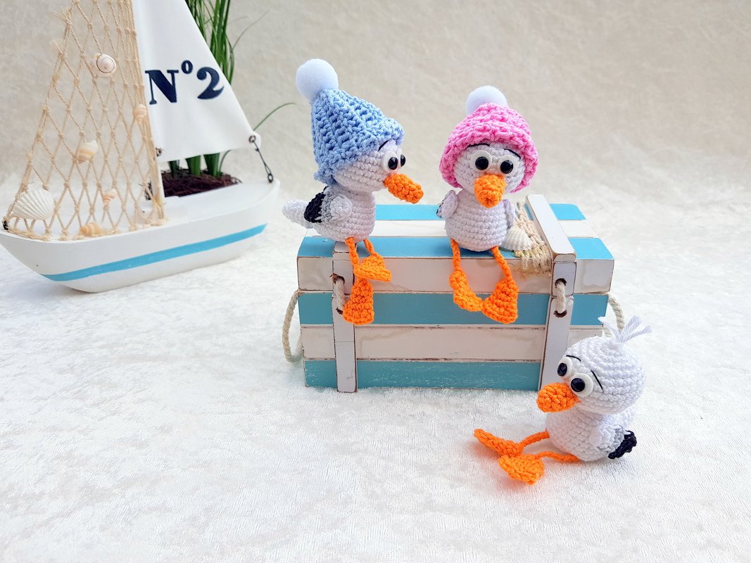 Handgehäkelte Amigurumi-Vögel mit Pastellmützen und orangen Schnäbeln auf einer gestreiften Holzbox neben einem Segelboot