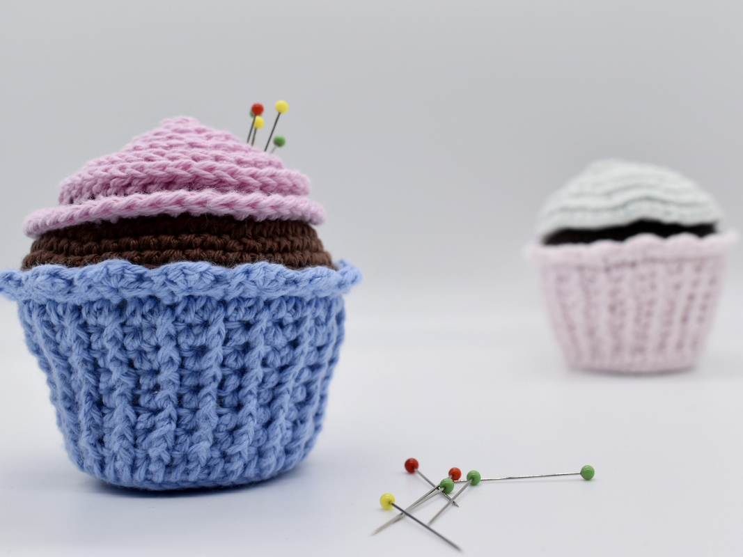 Gehäkeltes Cupcake-Nadelkissen mit Stecknadeln, weiteres Nadelkissen unscharf im Hintergrund