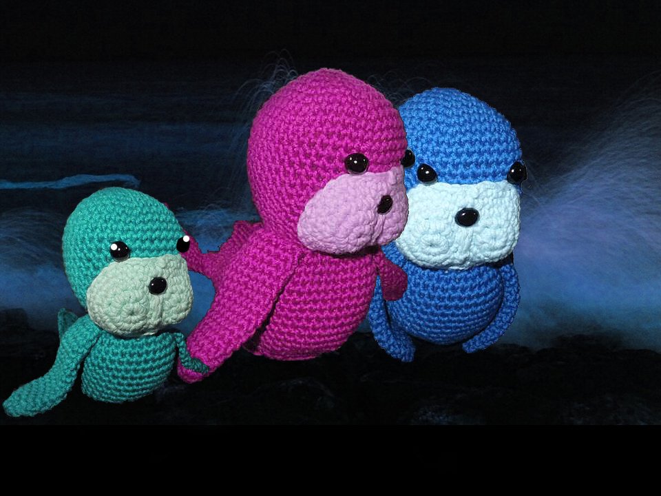 Drei gehäkelte Seeotter-Amigurumi (türkis, pink, blau) vor dunklem Hintergrund