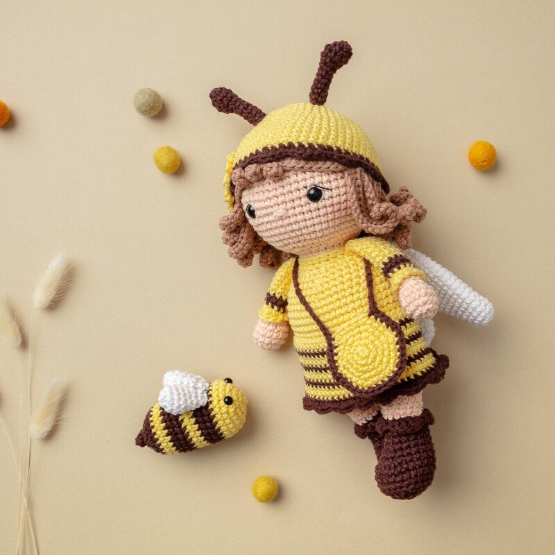 Gehäkelte Amigurumi-Puppe in gelb-braunem Bienenkostüm mit kleiner Biene auf beigem Hintergrund
