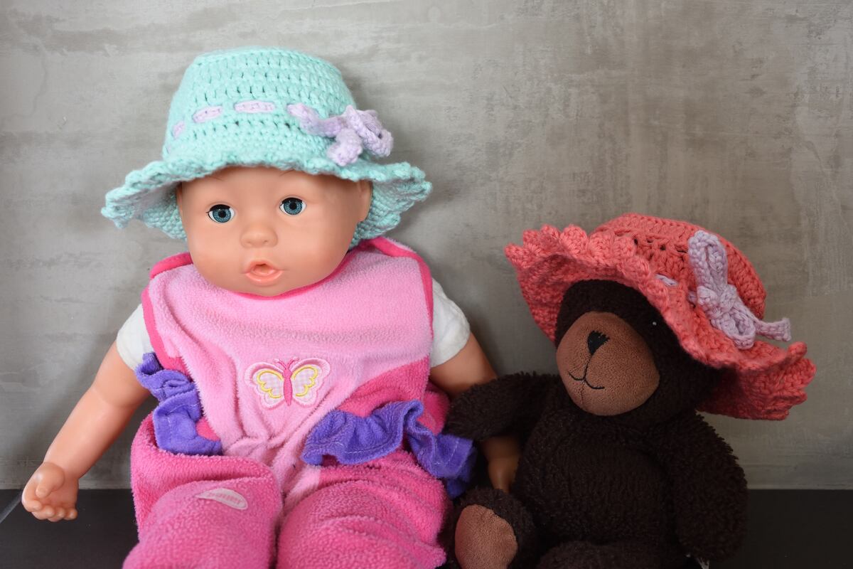 Mintgrüner gehäkelter Hut auf Puppe und korallfarbener gehäkelter Hut auf Teddybär, beide mit Rüschenrand und Schleife.