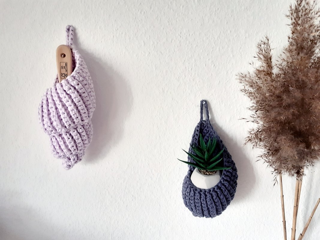 Zwei gehäkelte Wandutensilos in Lila und Dunkelblau, eines mit einem Gegenstand mit Holzgriff, das andere mit einer Sukkulente.