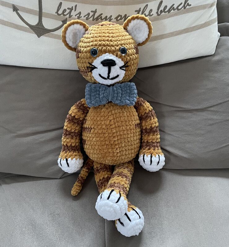 Gehäkelter Amigurumi-Tiger mit blau-grauer Fliege auf einem grauen Sofa.