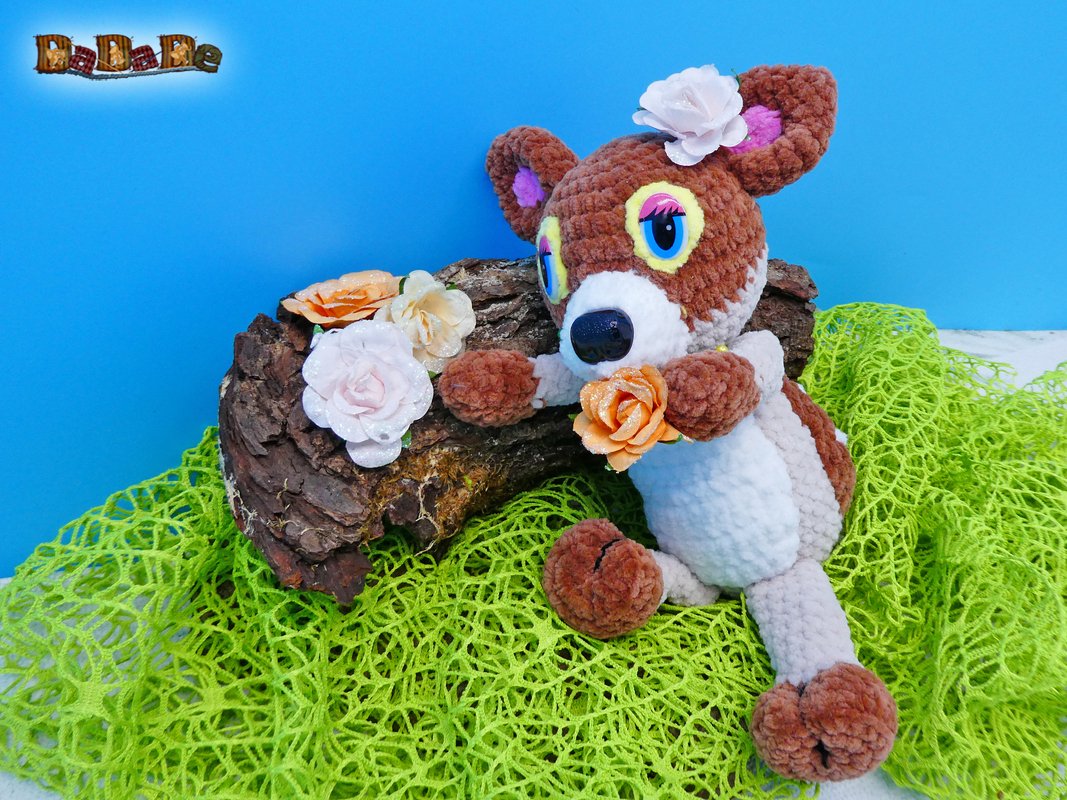 Gehäkeltes braun-weißes Rehkitz-Amigurumi mit Blume auf grünem Netzstoff vor blauem Hintergrund