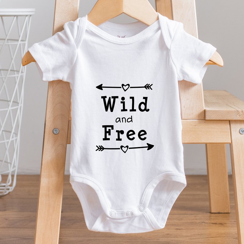 Weißer Babybody an einem Holzbügel mit schwarzem Schriftzug ‚Wild and Free‘, umrahmt von Pfeilen und Herzen.