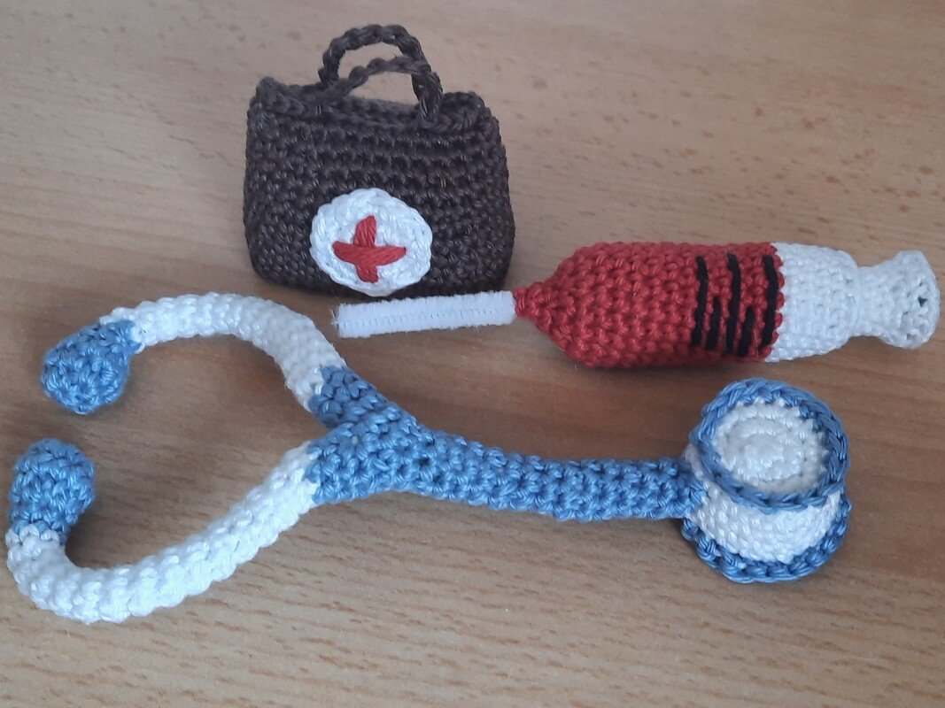 Gehäkeltes blau-weißes Stethoskop, rote gehäkelte Spritze und braune Arzt-Tasche mit weißem Aufnäher und rotem Kreuz