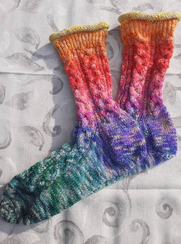 Paar handgestrickter Socken mit Farbverlauf von Grün über Violett zu Orange und Gelb, mit Zopfmuster und geripptem Bündchen, liegend auf hellem Stoff