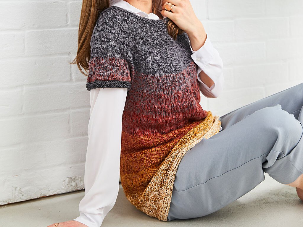 Person sitzt, trägt kurzärmelige gestrickte Tunika mit Farbverlauf von Grau zu Rot und Gold über weißer Bluse