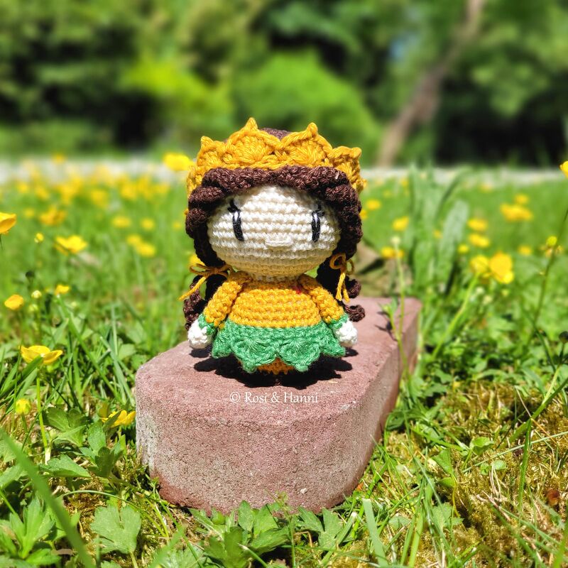 Gehäkelte Amigurumi-Puppe mit gelber Krone, gelbem Oberteil und grünem Rock auf einem Ziegelstein in einer Blumenwiese.
