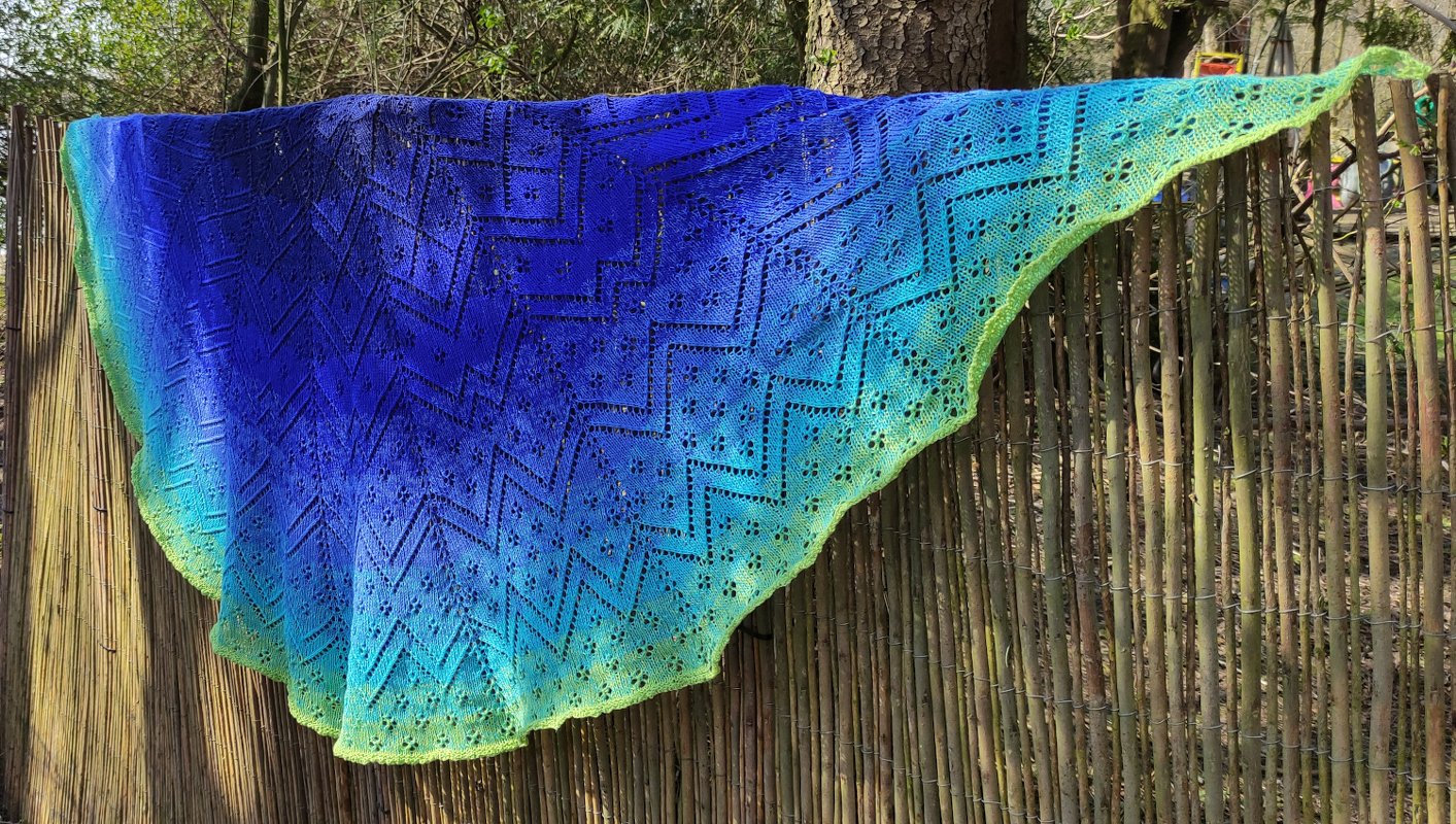 Halbrundes gestricktes Tuch mit Ajourmuster und Blau‑Grün‑Farbverlauf, aufgehängt an einem Zaun