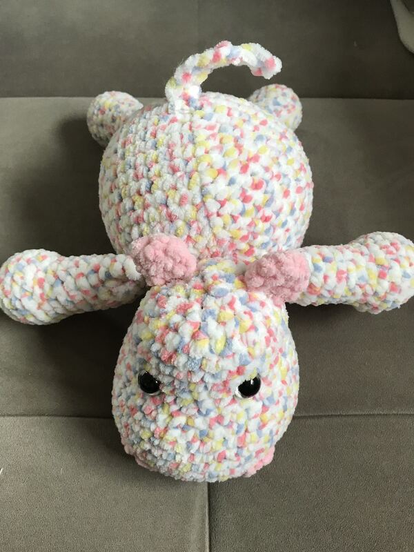 Gehäkeltes, flauschiges Amigurumi-Tier in pastellfarbenen Sprenkeln mit schwarzen Augen und rosa Ohren, liegend auf grauem Stoff