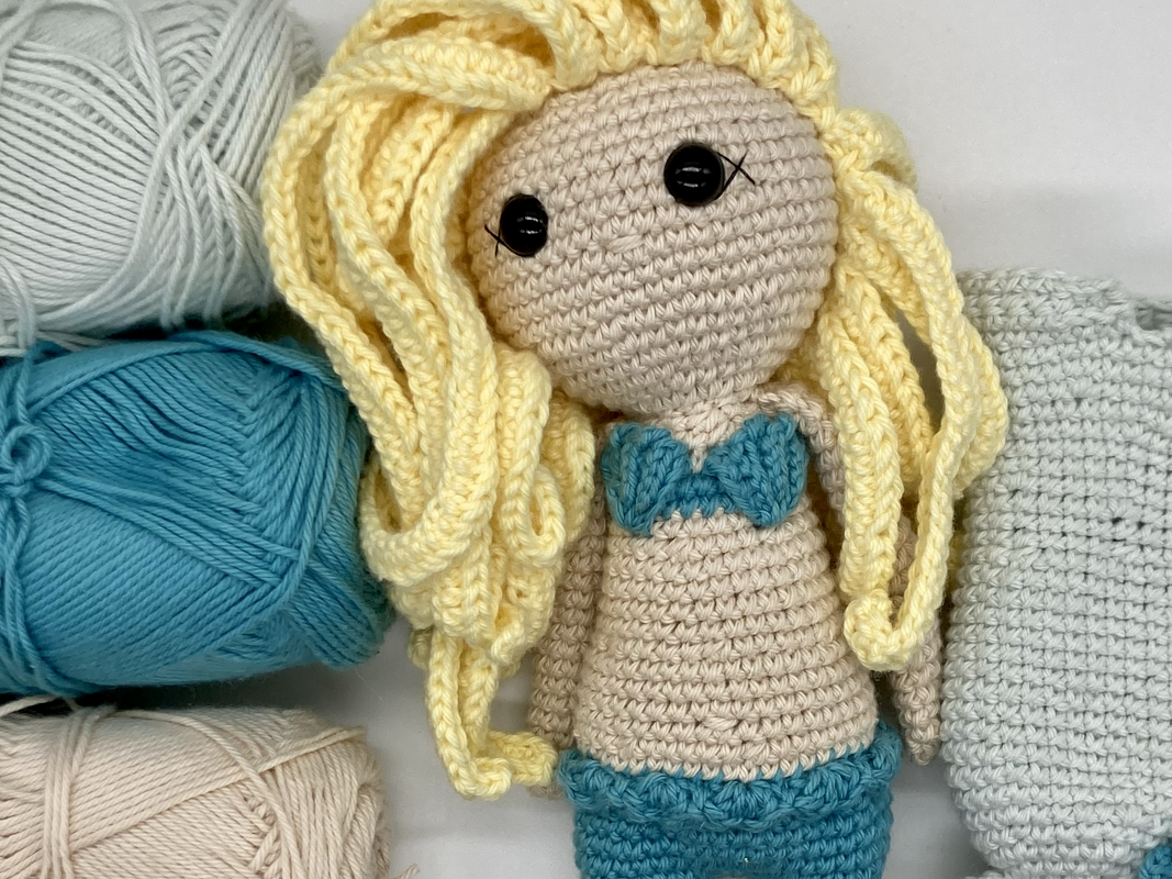 Gehäkelte Amigurumi-Puppe mit blondem Haar und türkisfarbenem Bikini, umgeben von Garnknäueln