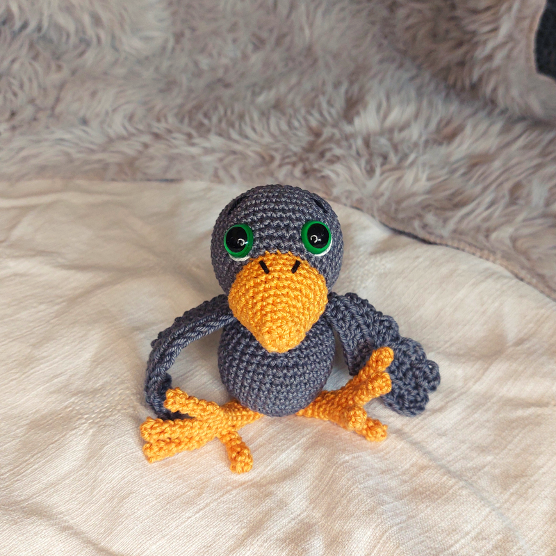 Hand-crocheted gray amigurumi bird with orange beak and feet and green eyes sitting on fabric