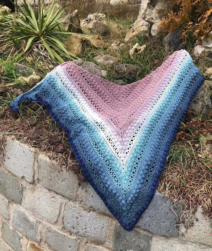 Gehäkeltes Dreieckstuch mit Farbverlauf in blau, türkis, weiß und rosa, auf einer Steinmauer und Gras drapiert.