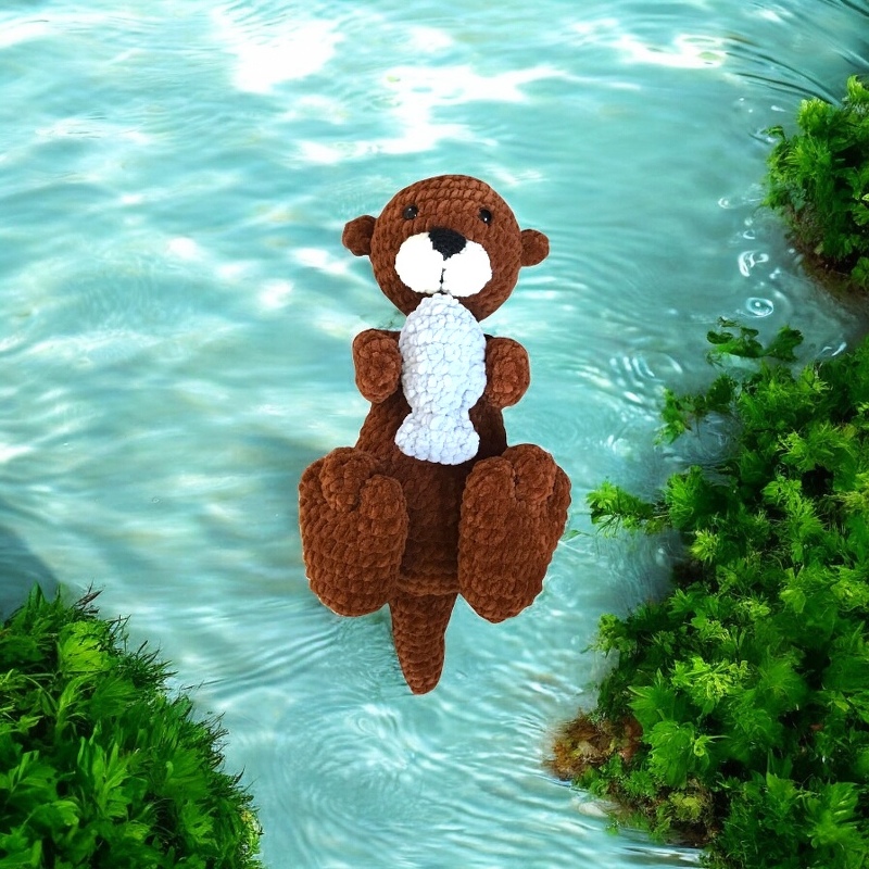 Braunes gehäkeltes Otter-Amigurumi, hält weißen Fisch vor blau-grünem Wasserhintergrund