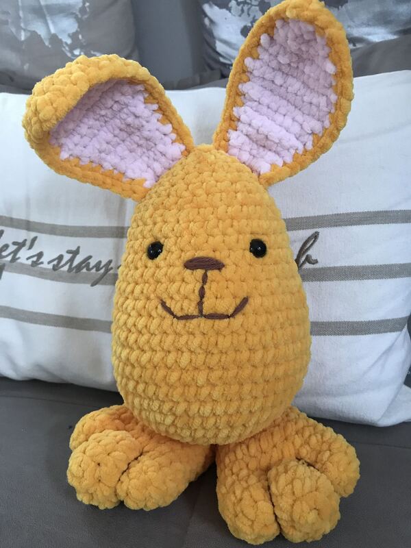 Gehäkelter gelber Hase mit rosa Ohren, schwarzen Augen und gestickter Nase, sitzend vor einem Kissen auf einer Couch.