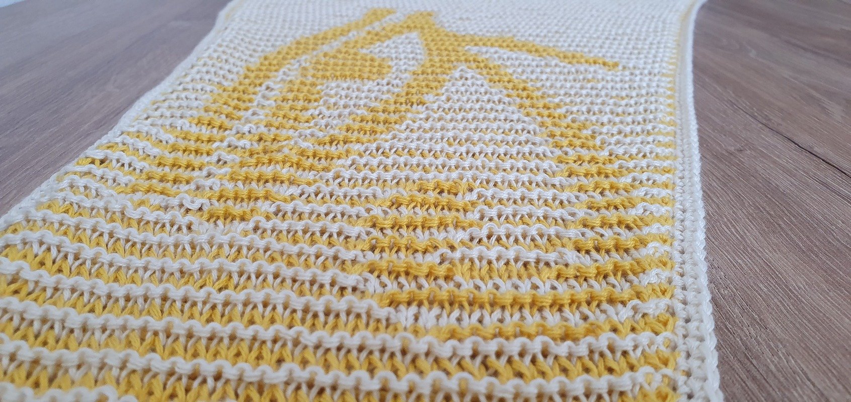 Cremeweißes und gelbes gestricktes Motivtuch mit Streifen und zentralem Sonnenmotiv auf Holzoberfläche.