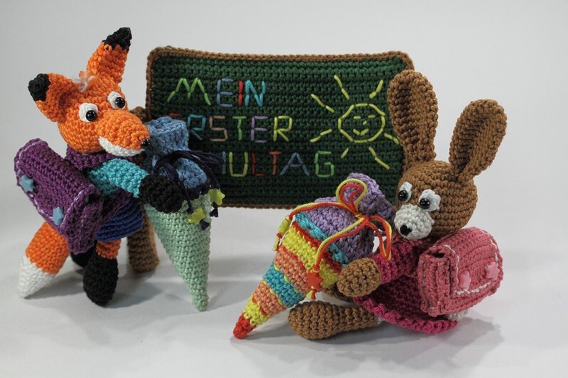 Gehäkelter Fuchs und Hase mit Schultüten und Schulranzen vor gehäkelter Tafel "Mein erster Schultag".