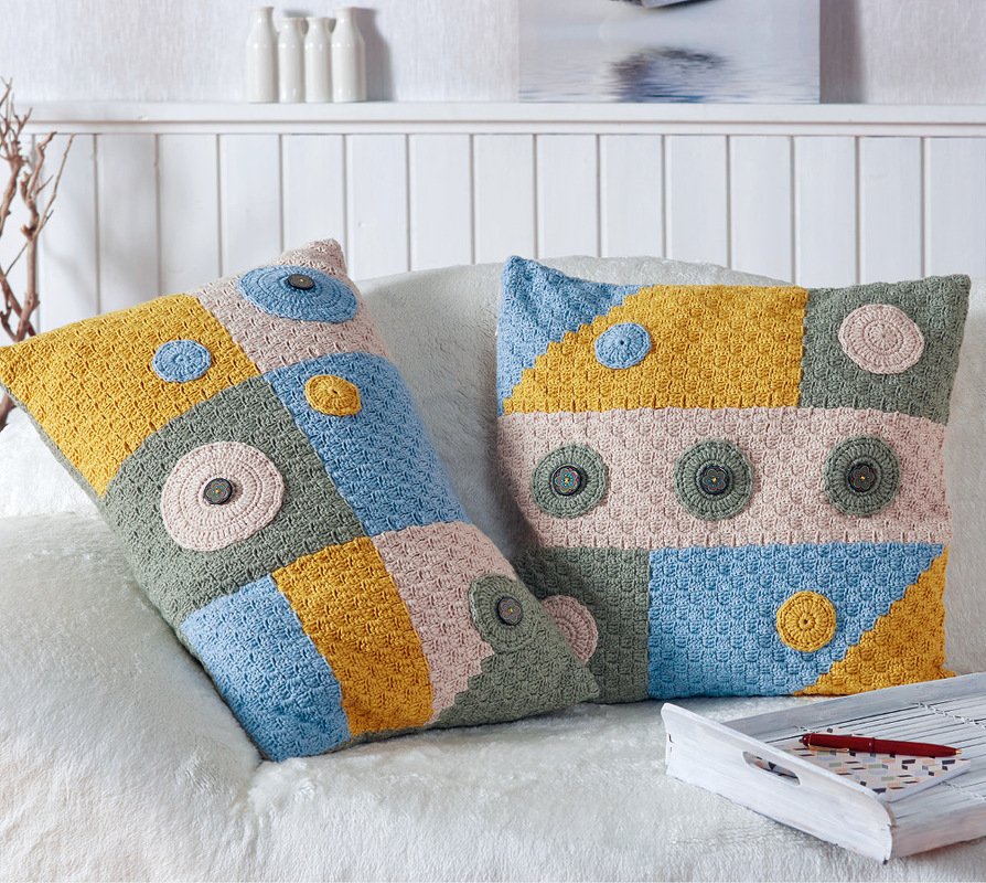 Zwei gehäkelte Patchwork-Kissenbezüge in Gelb, Blau, Rosa und Grün auf hellem Sofa