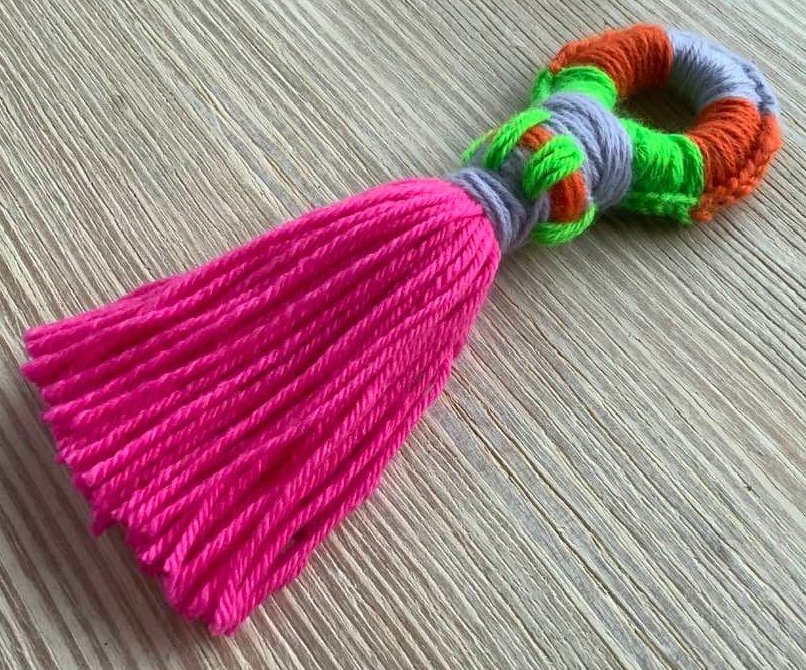 Gehäkelte Garnquaste mit pinken Fransen und mehrfarbigem Häkelring auf hellem Holzuntergrund.