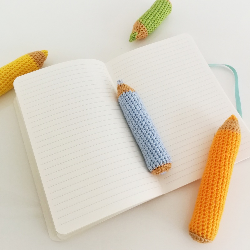 Mehrere gehäkelte Buntstifte in Gelb, Blau, Orange und Grün auf einem aufgeklappten linierten Notizbuch