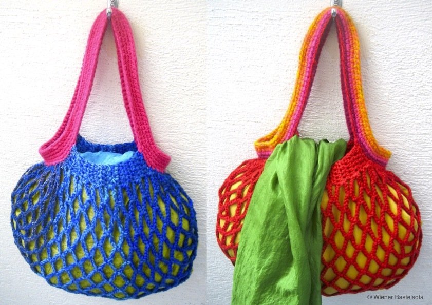 Zwei gehäkelte Netztaschen in Blau und Rot mit farbigen Henkeln, an einer hellen Wand hängend; gelber Inhalt sichtbar, grünes Tuch in der roten Tasche.