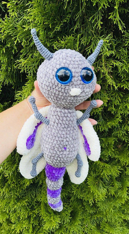 Gehäkelte Amigurumi-Motte in Grau, Weiß und Violett mit großen blauen Augen vor grünem Hintergrund