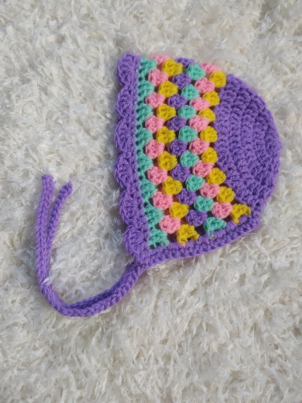 Lavender crocheted baby bonnet with pastel granny-stitch panel, scalloped edge and braided ties on a white textured background