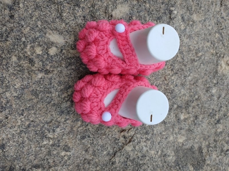 Pair of pink crocheted baby shoes with white button straps on a stone surface.