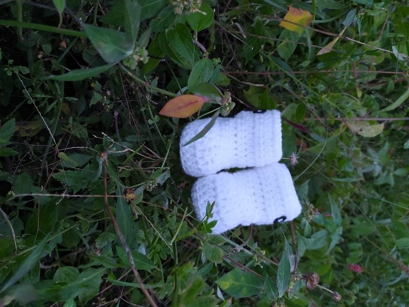 Pair of white crocheted baby booties with dark button accents on grass and leaves.