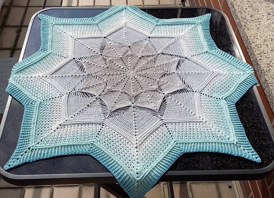 Star-shaped crocheted doily with gray-to-teal ombre on a dark table.